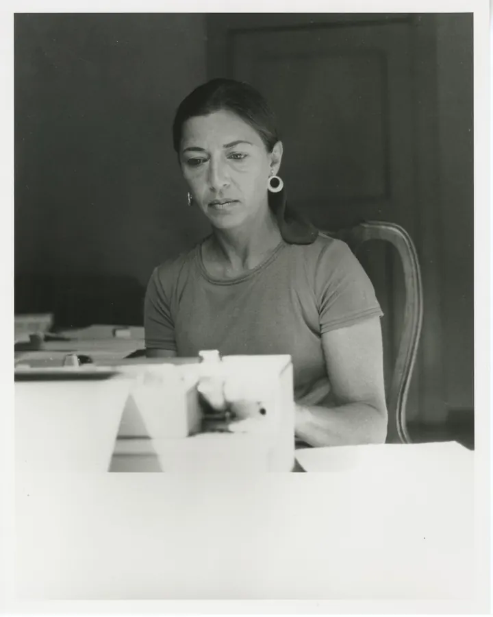 Femme aux cheveux foncés assise à une table, portant un t-shirt et des boucles d'oreilles rondes, devant une machine à écrire dans une pièce sombre.