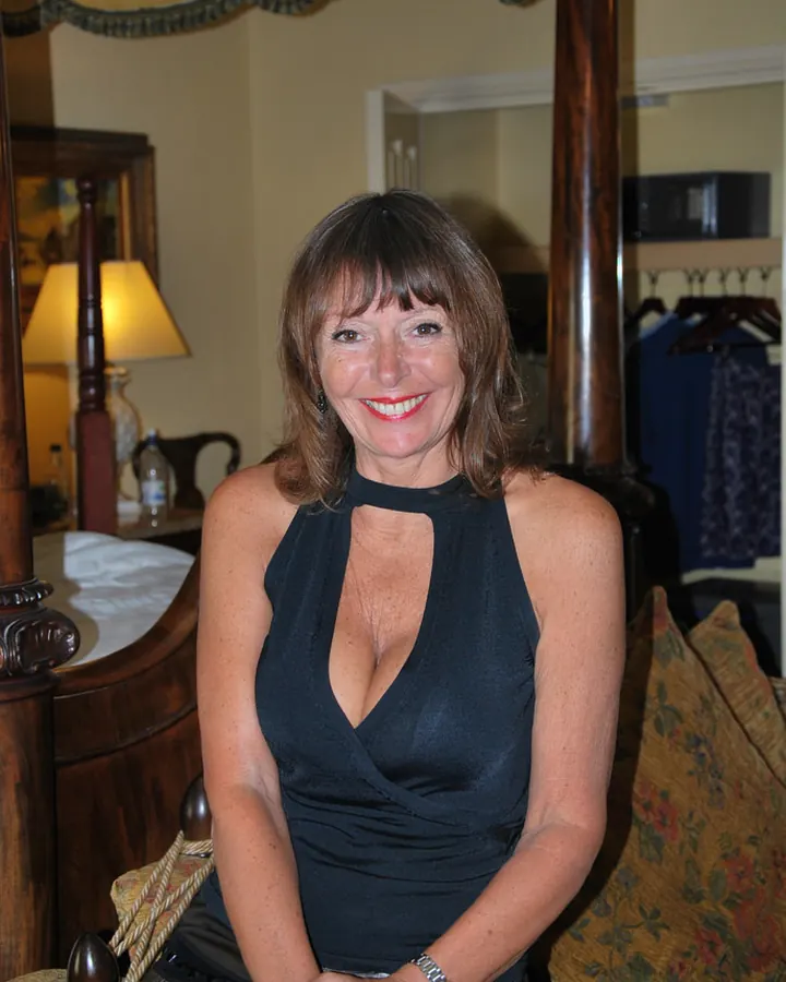 Femme aux cheveux châtains, souriante, portant un haut noir et une jupe grise, assise sur une chaise dans une chambre avec une lampe allumée.
