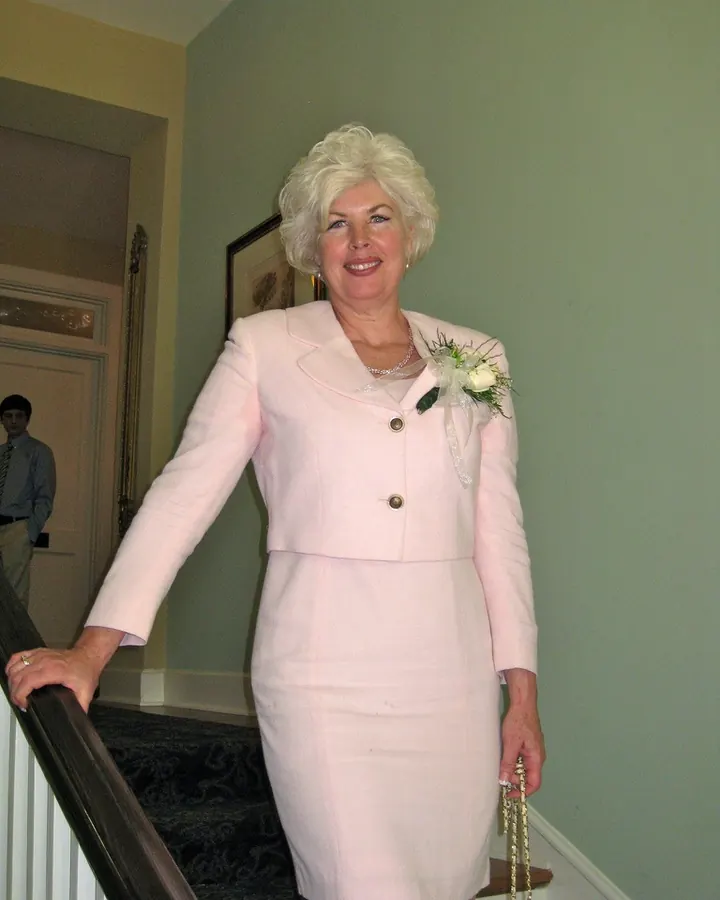 Femme aux cheveux platine, souriante, portant une veste rose pâle et une jupe assortie, tenant un bouquet de fleurs, debout sur un escalier à l'intérieur d'une maison.