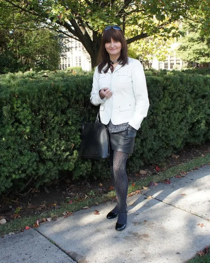 Femme aux cheveux bruns-noirs, peau blanche, poitrine petite, en veste blanche et jupe en cuir, debout sur un trottoir avec des buissons.