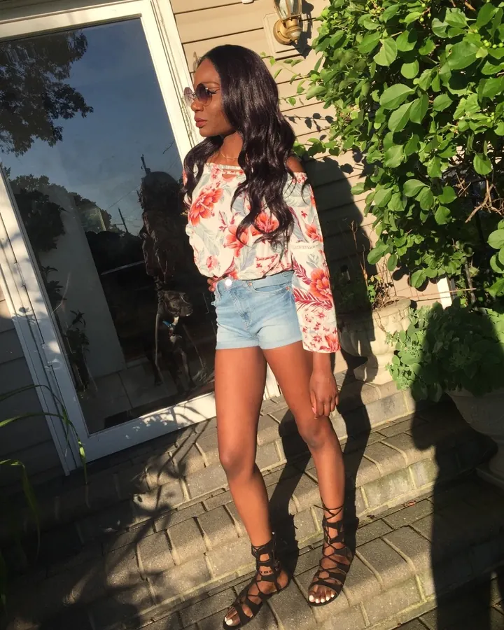 Femme aux cheveux bruns-noirs, peau olive, en chemisier fleuri et shorts en jean, debout sur un perron ensoleillé avec des plantes vertes.