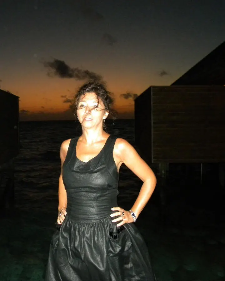 Femme aux cheveux bruns-noirs, peau moyenne, en robe noire, debout sur un pont au crépuscule, mains sur les hanches.