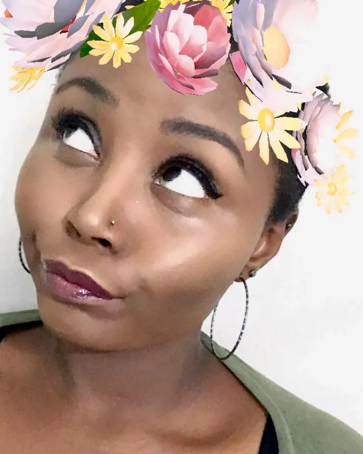 Femme aux cheveux bruns-noirs, peau noire, yeux noirs, en chemisier floral et shorts en jean, debout sur un perron avec des plantes.
