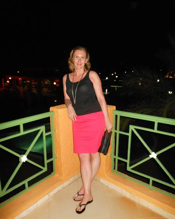 Femme blonde en robe noire, souriante, debout sur un balcon la nuit, tenant un sac noir.