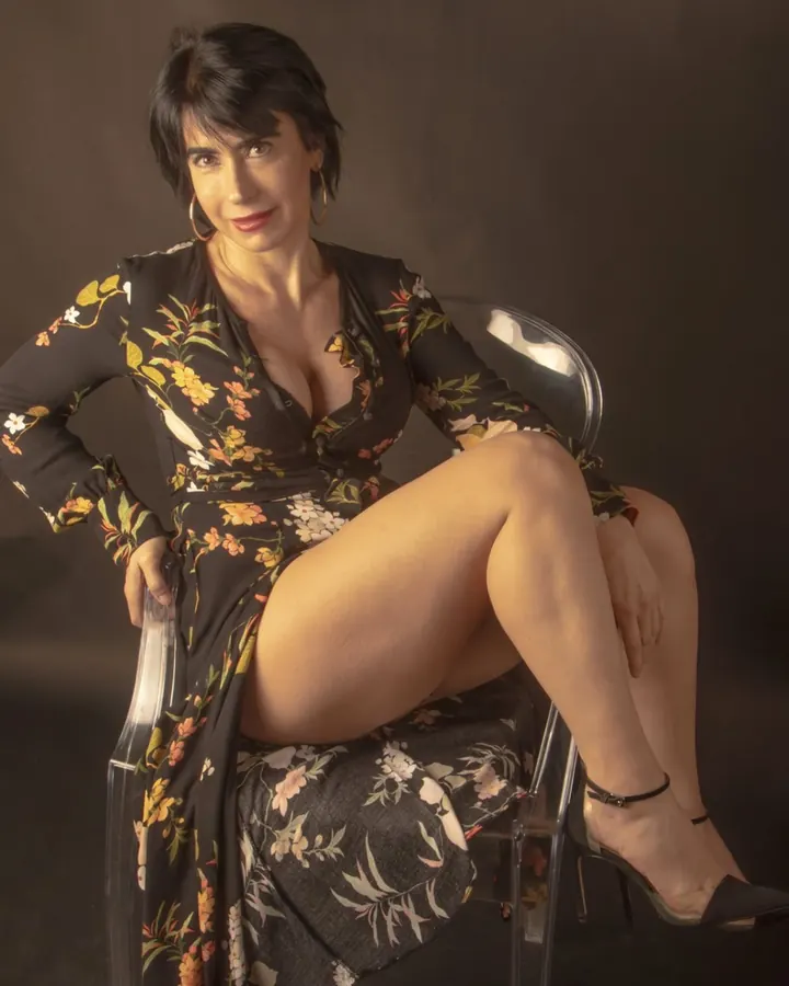 Femme aux cheveux noirs en robe fleurie noire, assise sur une chaise transparente, les jambes croisées, regardant la caméra.
