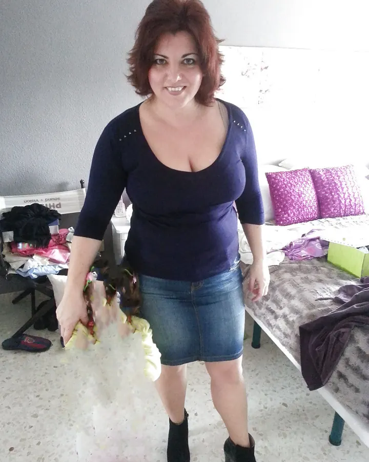Femme aux cheveux bruns en haut bleu marine et jupe en jean, debout dans une chambre, tenant une poupée.