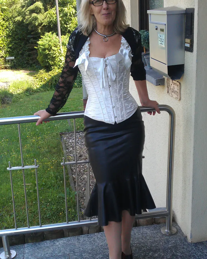 Femme blonde aux yeux marrons en corset blanc et jupe noire, posant sur un balcon