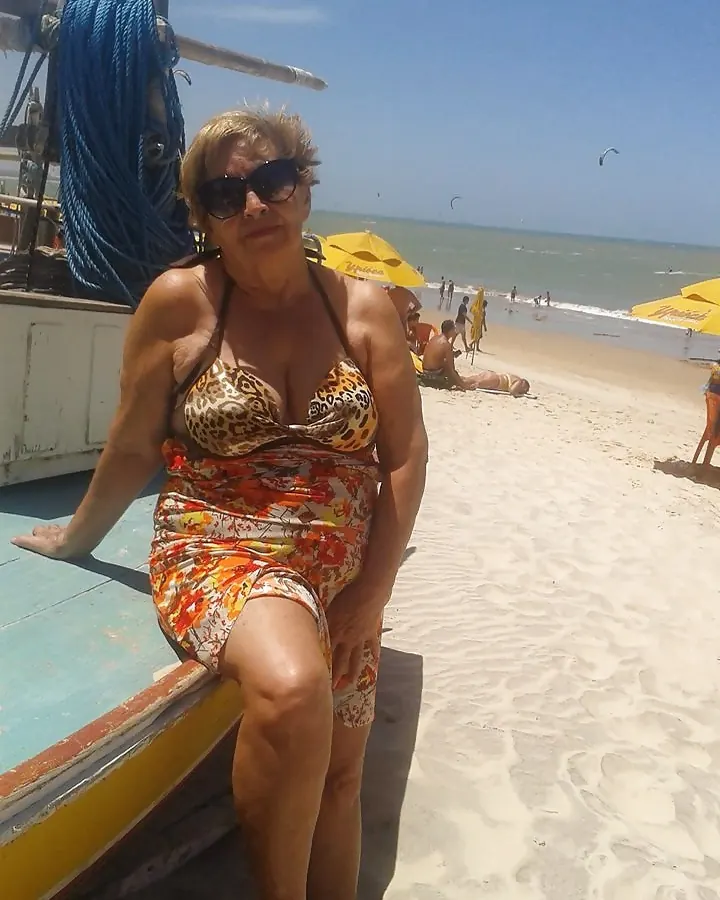 Femme aux cheveux blonds, en bikini léopard, pose sur un bateau au bord d'une plage, lunettes de soleil, regard direct.