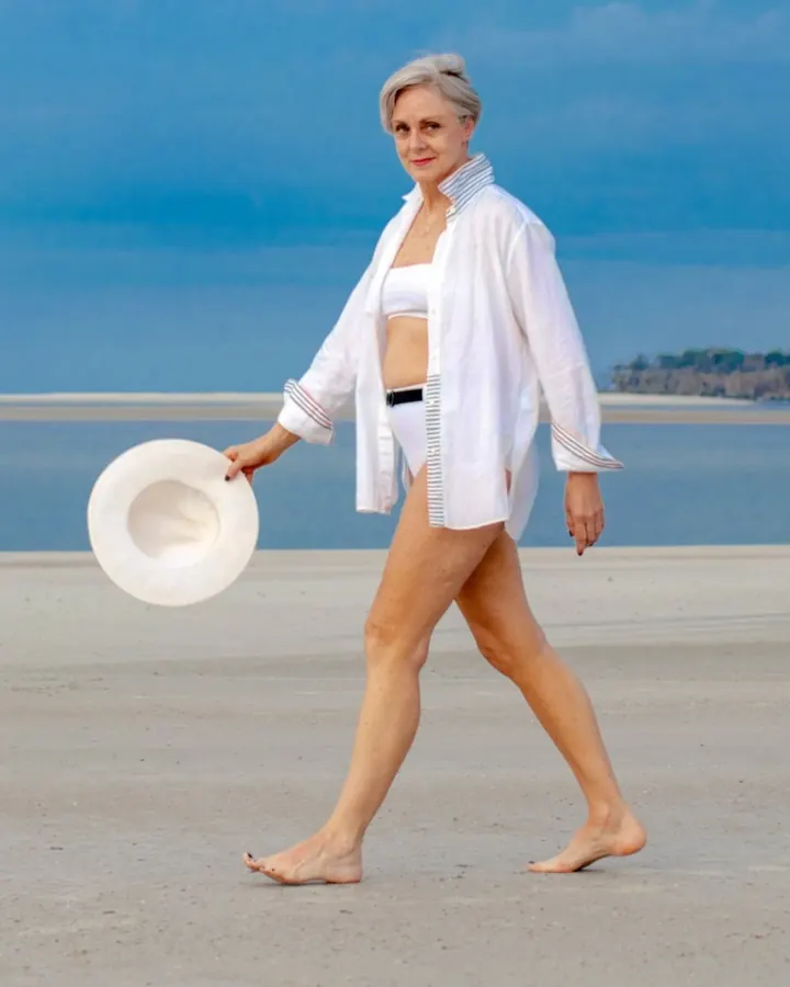 Femme blonde, en bikini blanc, portant une chemise blanche ouverte, marchant pieds nus sur une plage avec un chapeau blanc à la main