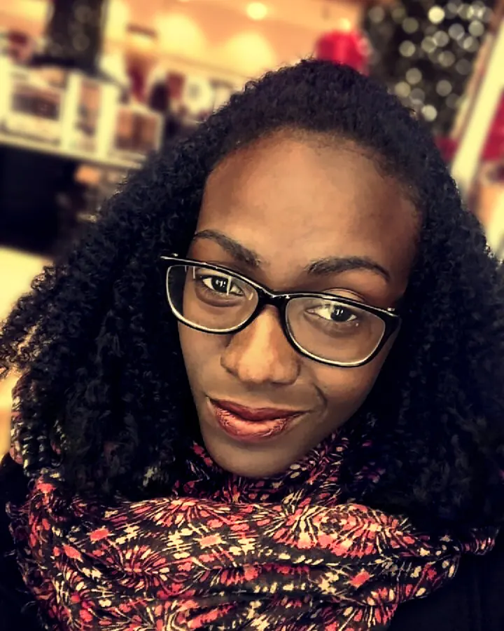 Femme aux cheveux noirs bouclés, portant des lunettes et un foulard coloré, souriant à l'appareil photo.