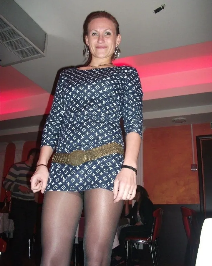 Femme aux cheveux châtains, portant une robe à motifs Louis Vuitton, une ceinture dorée et des collants, souriant à la caméra dans un lieu intérieur.