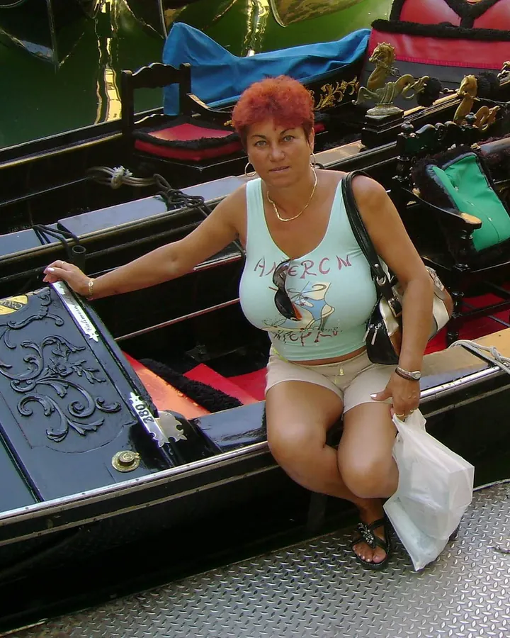Femme aux cheveux roux, vêtue d'un débardeur turquoise et d'un short beige, assise sur un bateau décoré, tenant un sac en plastique.