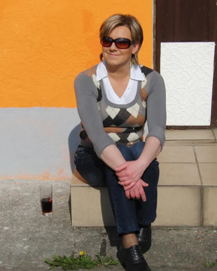 Femme blonde portant un haut gris et un pantalon bleu, assise sur un marchepied, les mains croisées, avec lunettes de soleil, dans un extérieur avec mur orange et verre de boisson.