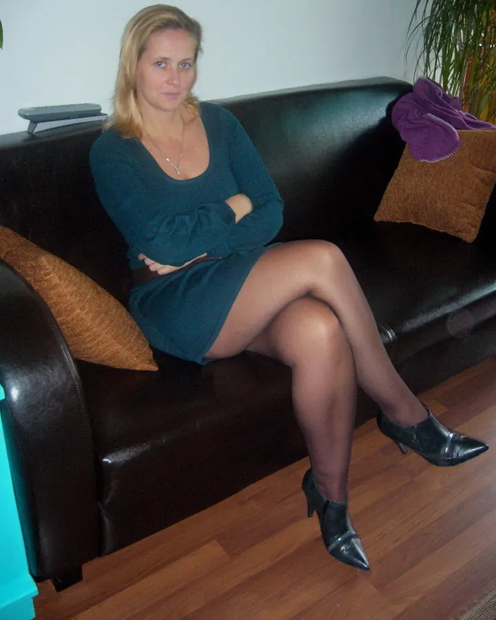 Femme blonde assise sur un canapé noir, vêtue d'une robe verte, les bras croisés, portant des collants noirs et des escarpins noirs pointus, dans un salon avec plantes et coussins colorés.