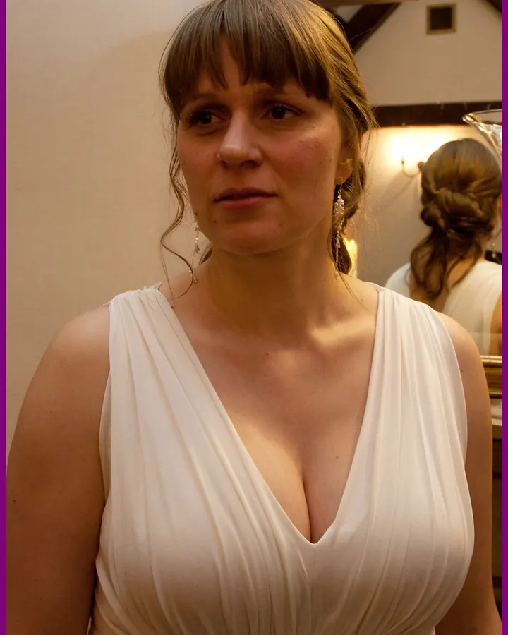 Femme aux cheveux blonds portant une robe blanche à décolleté plongeant, regardant de côté, dans un intérieur avec lumière chaude et miroir en arrière-plan.
