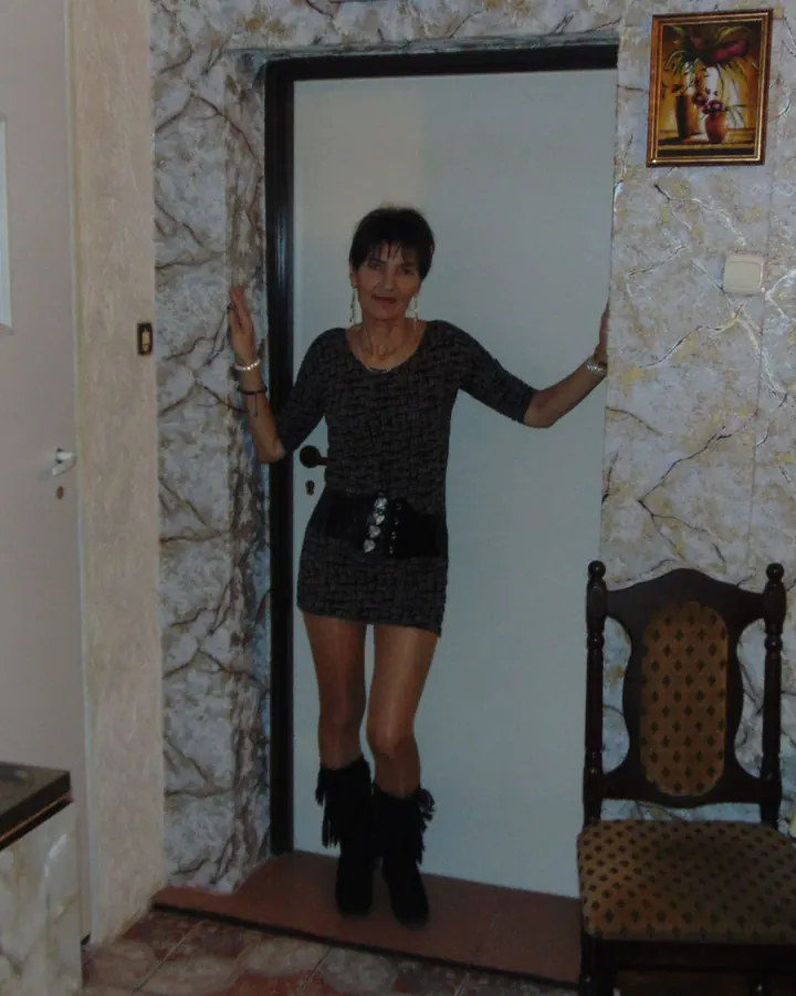 Femme aux cheveux bruns-noirs portant une robe noire, allongée sur un lit blanc, les jambes croisées, portant des collants noirs et des escarpins noirs, dans une chambre avec lampe et téléphone.