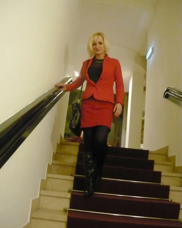 Femme blonde portant une robe marron, assise sur un escalier, les mains sur les barrières, portant des collants noirs et des bottes noires, dans un couloir avec tapis rouge.