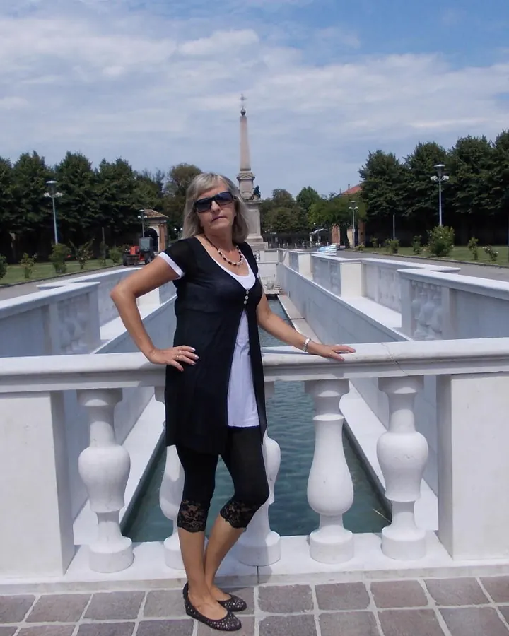 Femme aux cheveux gris en tenue noire, debout sur un pont, regard sérieux, lunettes de soleil.