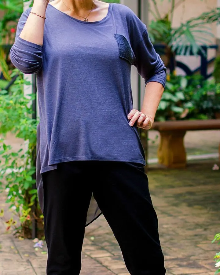 Femme souriante, aux cheveux courts gris-bleu, portant des lunettes rouges, un haut bleu et un pantalon noir, debout sur un patio verdoyant.