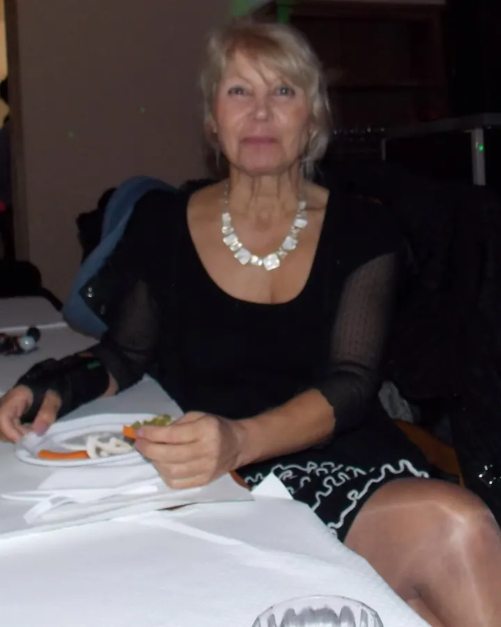 Femme aux cheveux blonds courts, souriante, portant une robe violette sans manches, debout devant une cheminée décorée de Noël.