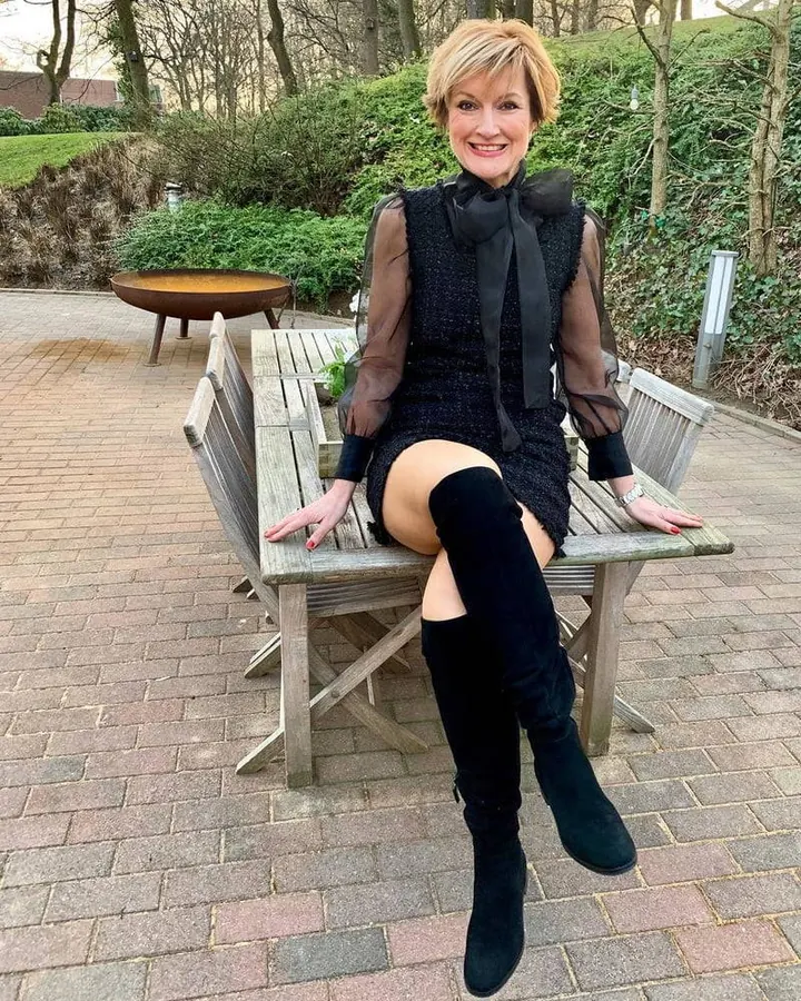 Femme aux cheveux blonds courts, souriante, portant un haut noir et une jupe rouge, assise sur un canapé rouge.