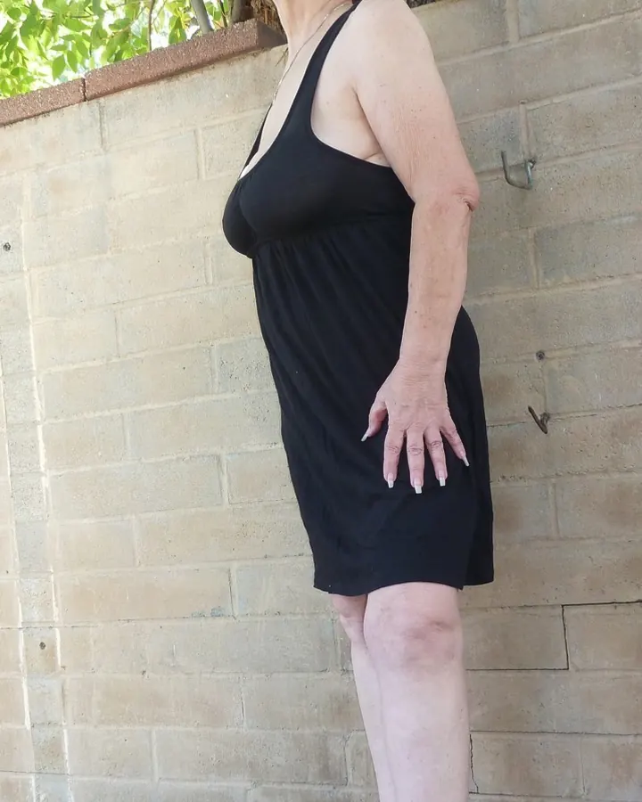 Femme aux cheveux roux portant une robe noire, debout contre un mur de briques en extérieur.
