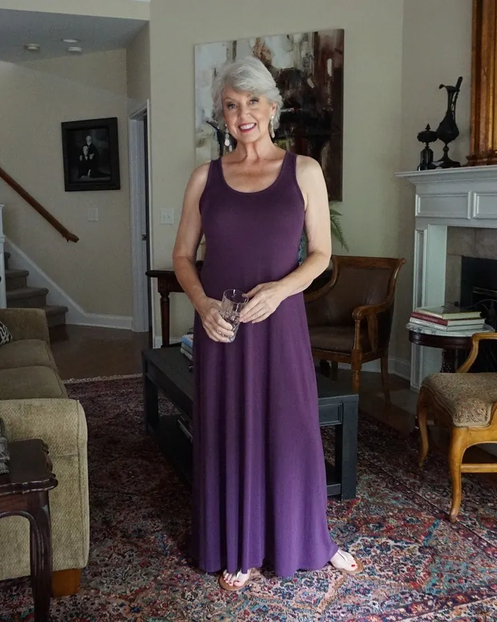 Femme aux cheveux gris clair, souriante, en robe violette longue, tenant un verre dans un salon élégant
