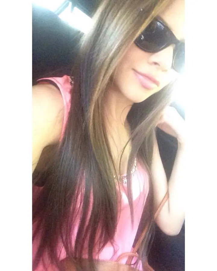 Femme aux cheveux longs, portant des lunettes de soleil et un haut rose, assise dans une voiture.