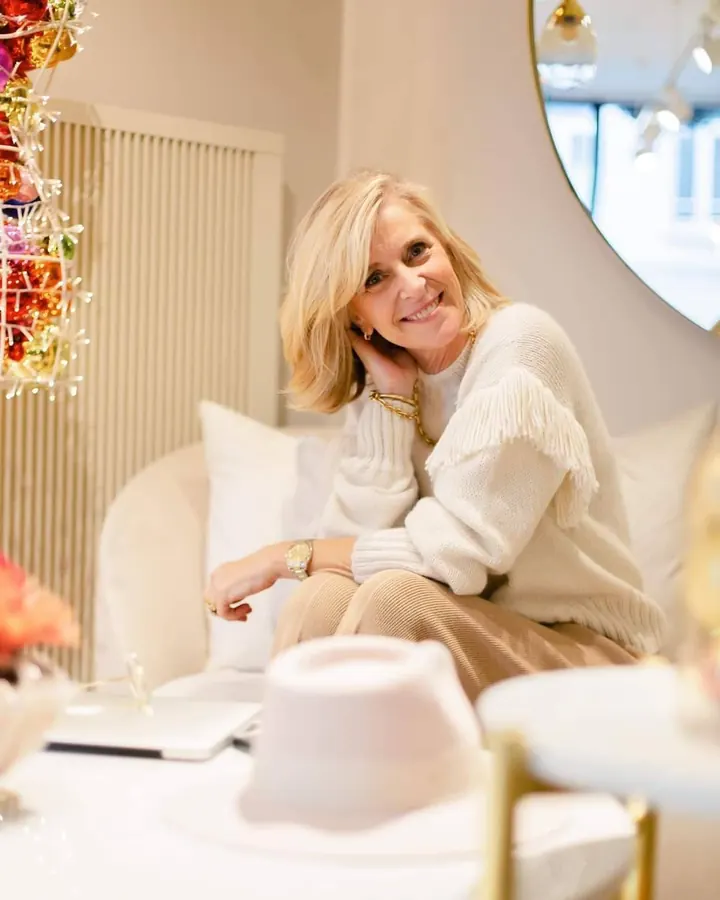 Femme aux cheveux blonds portant un pull blanc à franges, assise sur un canapé blanc, près d'un sapin décoré