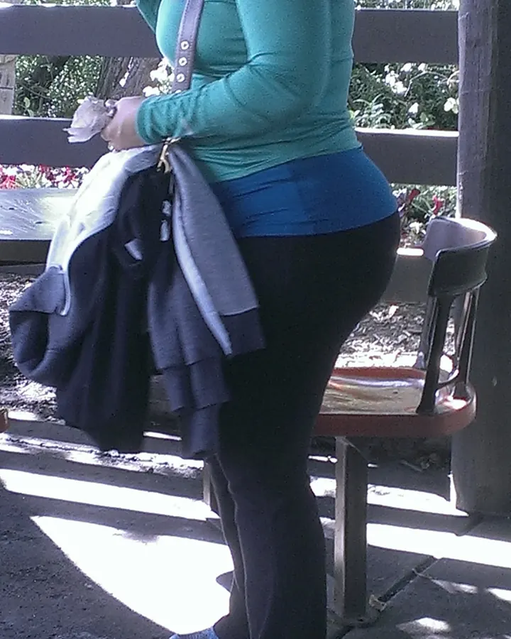 Vue arrière d'une femme en tenue verte et bleue, tenant un sac avec un pull gris, debout près d'un banc en bois