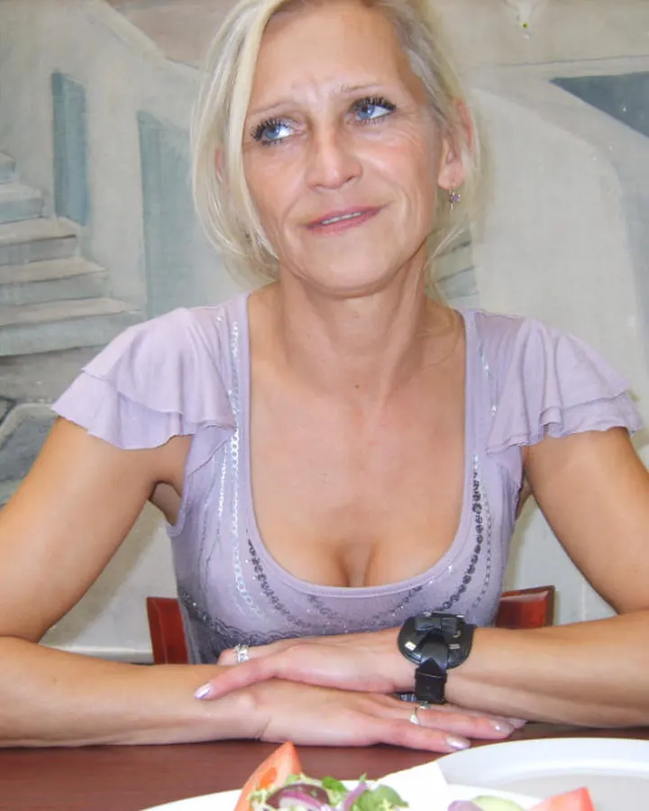 Femme aux cheveux blonds platine courts, souriante, en haut lilas, assise à table avec un verre de vin blanc.