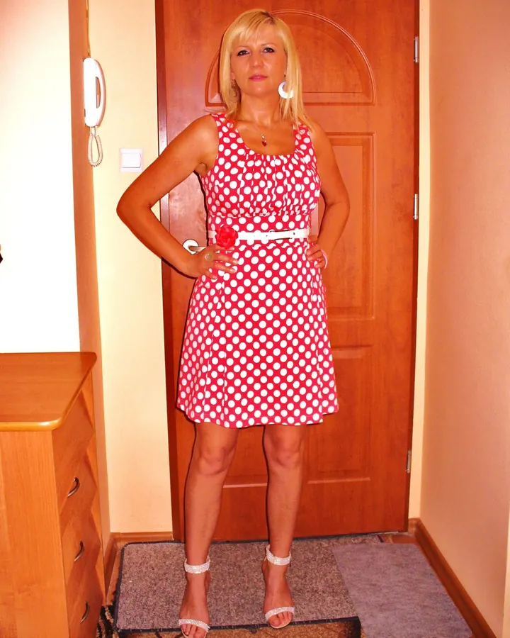 Femme aux cheveux blonds courts, souriante, en robe rouge à pois blancs, debout devant une porte en bois.
