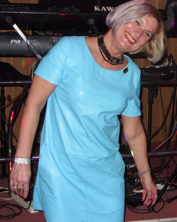 Femme aux cheveux blonds courts, souriante, en tenue noire en cuir, bottes hautes, debout dans une cuisine.