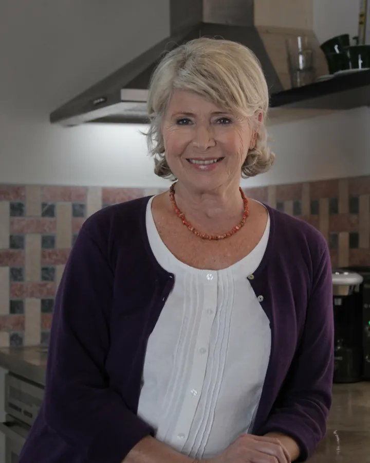 Femme aux cheveux blonds courts, souriante, en pull violet sur un haut blanc dans une cuisine moderne.
